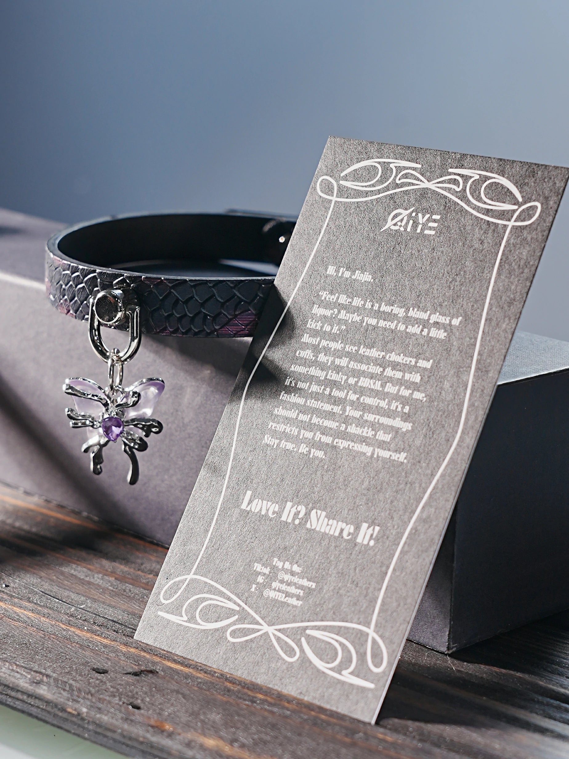 QIYE_Leather_Banner_Collar_picture: Scene image of a leather collar with a snake - skin texture and a butterfly decorative charm, placed alongside a grey card with decorative text.