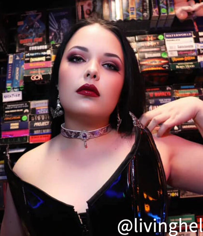 The Tear Of Ames-Violet Jewelry, a striking violet choker with a dangling gem, adorns a model wearing a glossy black outfit. The scene is set against a backdrop of colorful VHS tapes, highlighting the choker's elegant design.