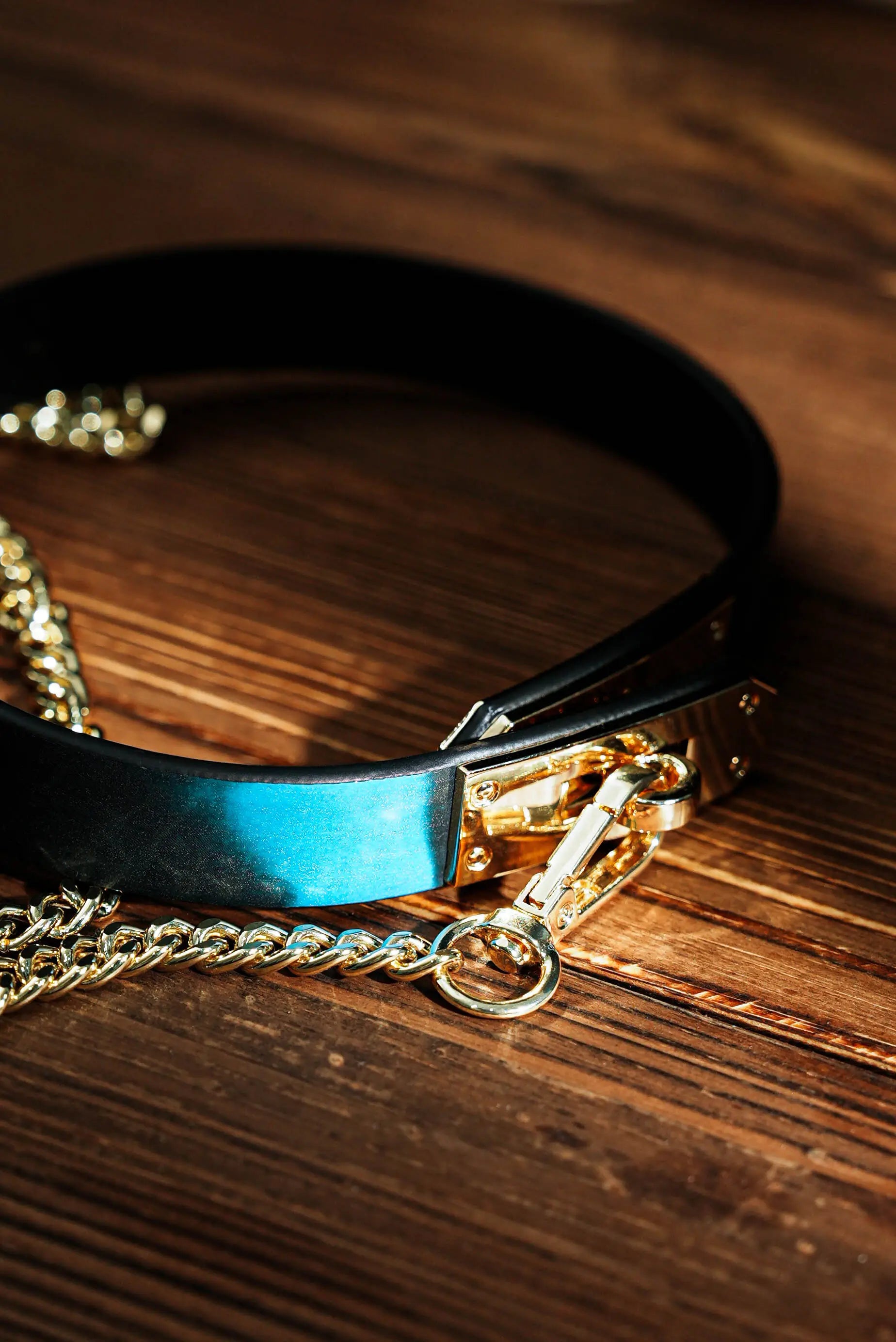 QIYE_Leathers_Heat - Reactive_Collars_Comet_color: A heat - reactive collar placed on a wooden background, featuring black leather with a blue heat - reactive panel, and shiny gold - toned metal chains and clasps.