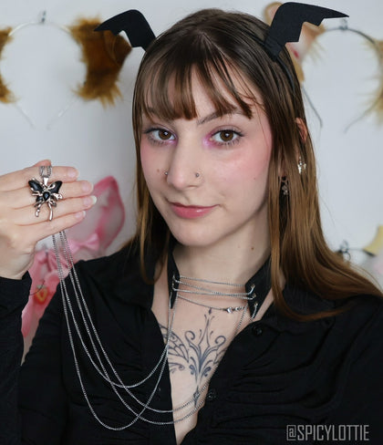 A model showcases the Roxey-Leather Choker With Butterfly, prominently displaying the intricate butterfly accessory in hand. The choker's delicate silver chains cascade beautifully over a black top, set against a whimsical background with animal ears and pastel accents.
