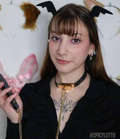 A model wears the Glimmer-Heat Reactive Choker, featuring a sleek collar and a dangling gold chain, while holding up a matching wrist strap. The scene is set against a whimsical background with bat and animal ear decorations.